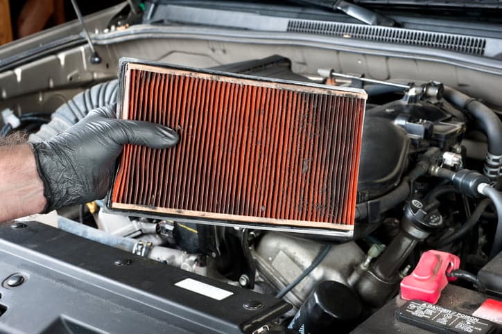 An auto mechanic wearing protective work gloves holds a dirty, clogged air filter over a car engine during general auto maintenance.